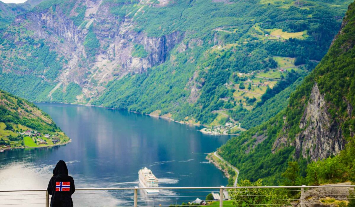 Tourist wearing norwegian flag clothing enjoying scenic view over fjord Geirangerfjorden from Flydalsjuvet viewpoint. Cruising vacation and travel.