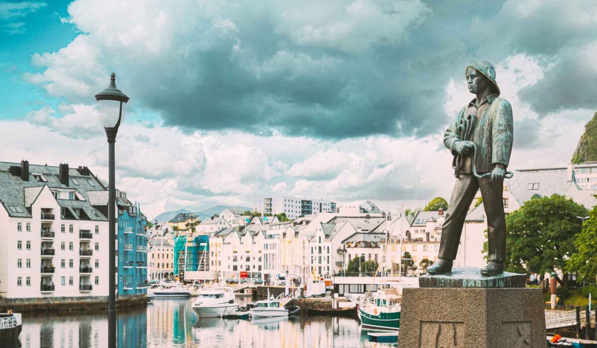 Alesund, Norway. Statue Of Young Sailor-fisher Boy On Brosundet Canal. Old Wooden Houses In Summer Day.