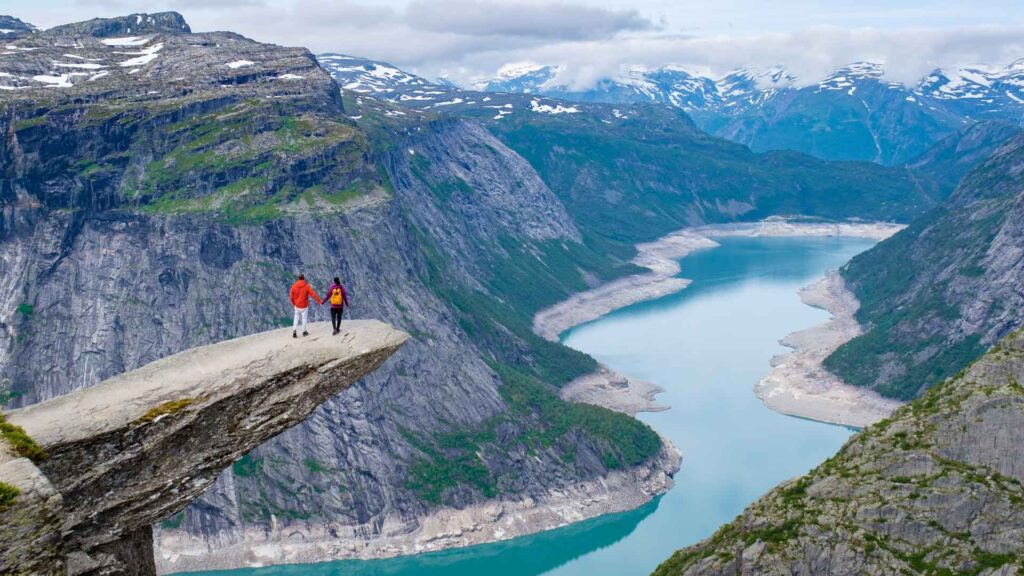 Two hikers stand on a rocky precipice overlooking a picturesque Norwegian fjord, with snow-capped mountains in the distance. Trolltunga, Norway,