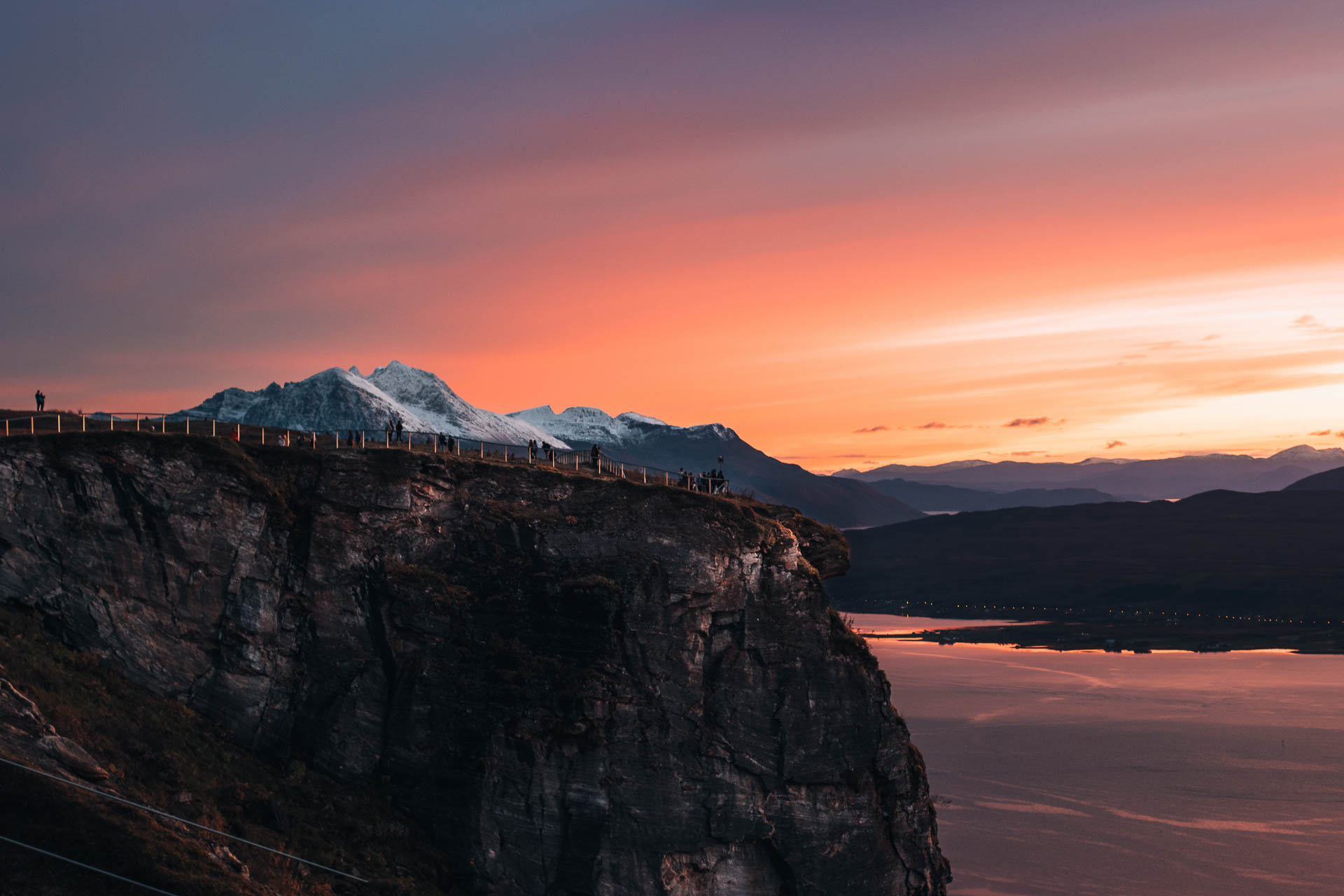 Tromsø Travel Guide 22 Cliff at Fjellheisen viewpoint in Tromsø, Norway, with a breathtaking sunset and snow-capped mountains in the background.